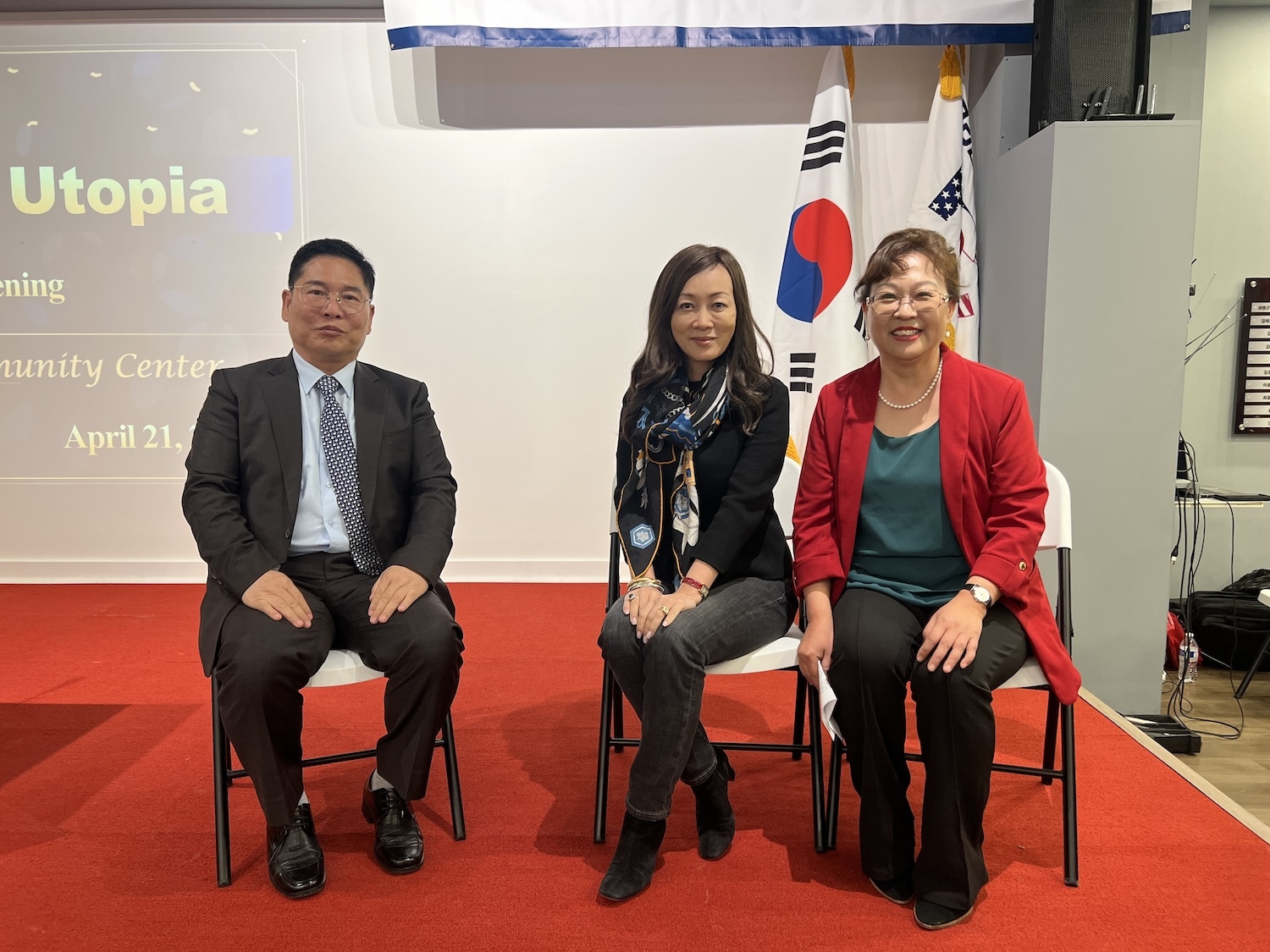 Three individuals are seated on white chairs in front of a presentation screen and a South Korean flag in a community center, ready to discuss the human rights crisis in North Korea as part of the Beyond Utopia initiative.