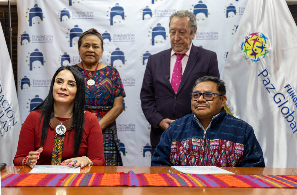 Four individuals, including Nobel Laureate Rigoberta Menchú, sit and stand in front of a backdrop with logos, in a formal setting. Two are seated at a table adorned with vibrant woven fabric, while the other two stand behind, symbolizing their commitment to Peace in Central America.