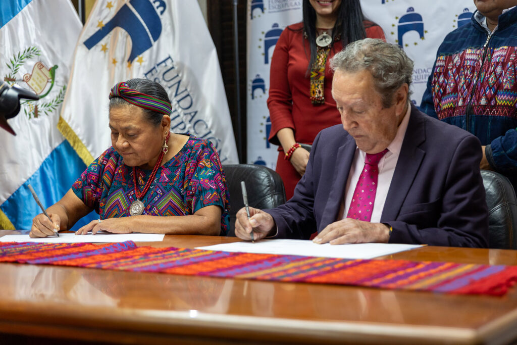 Two individuals sit at a table, signing documents. Colorful textiles adorn the table, reflecting a rich cultural heritage. Nobel Laureate Rigoberta Menchú stands nearby, observing the event. Guatemalan flags and a banner with logos in the background highlight this Cooperation Agreement for Peace.