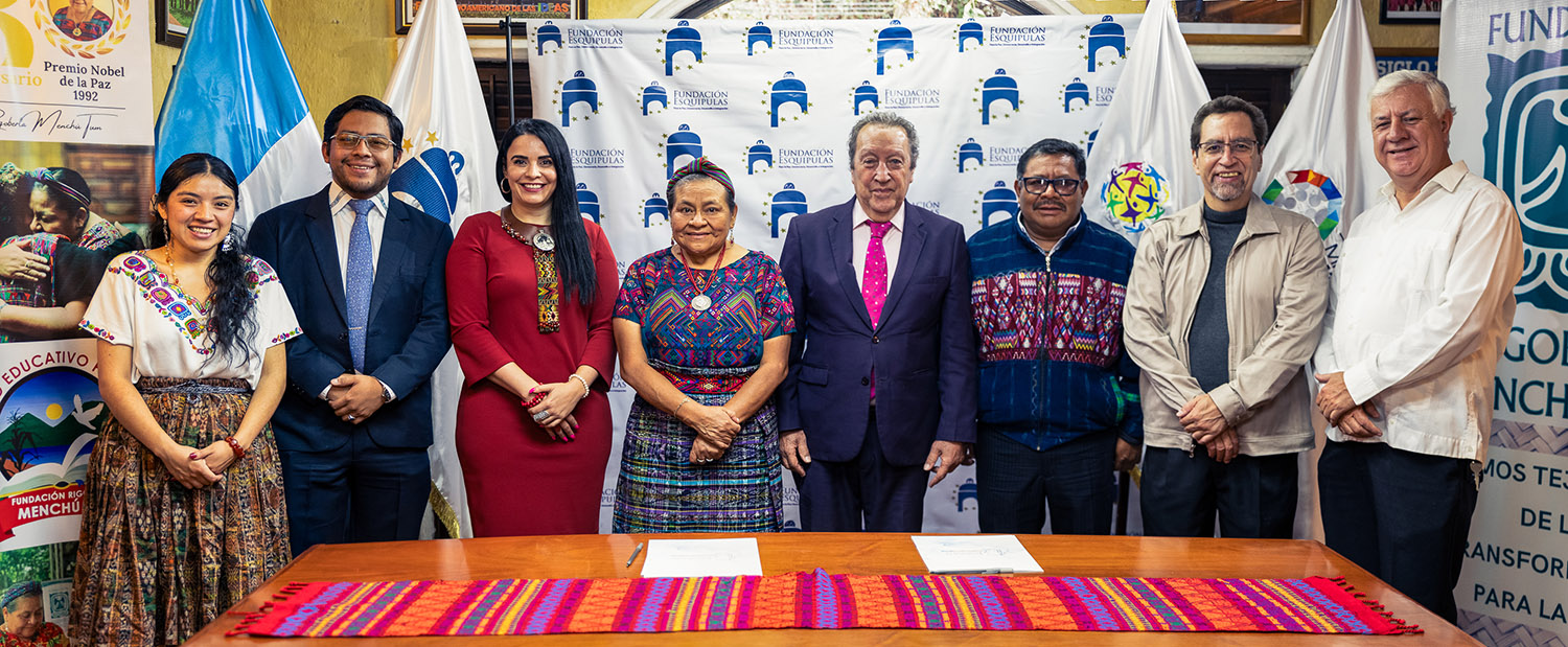 A group of eight people stands behind a table. They are posing for the photo in front of banners and flags, and some are wearing traditional clothing. Highlighting their commitment to regional stability, they’ve just signed a Cooperation Agreement for Peace in Central America.