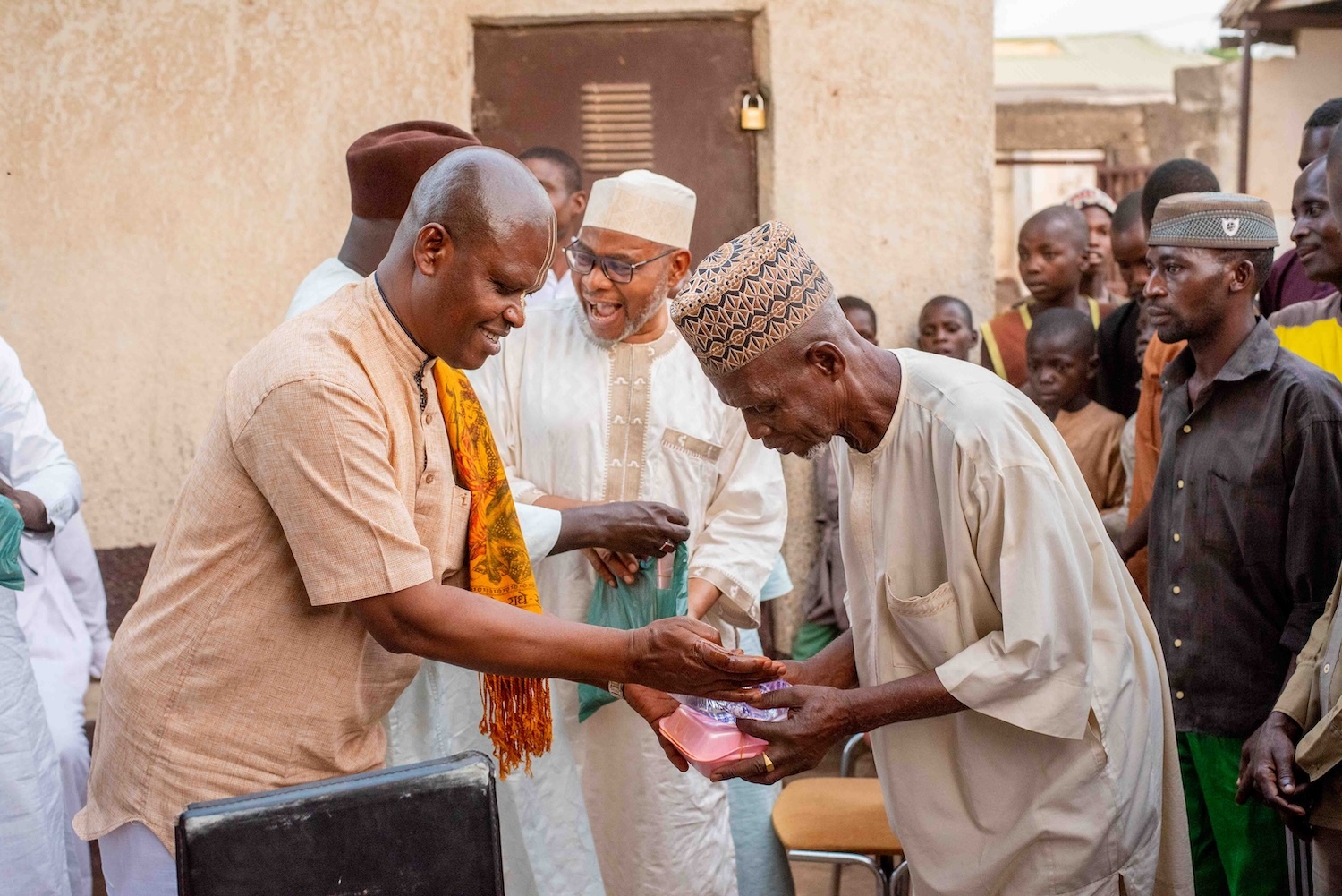 Two elderly men in traditional Nigerian attire exchanging a gift outdoors, surrounded by a group of onlookers.