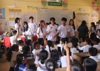 As part of the One Book One Love Campaign, a group of students in white shirts interact with younger children in a classroom decorated with educational posters and drawings. Several children raise their hands while seated at their desks, eager to participate.