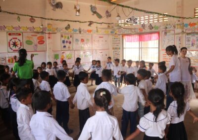A group of young students in Cambodia, dressed in uniforms, stands in a circle holding hands in a classroom decorated with colorful educational posters and hanging crafts from the "One Book One Love" campaign.