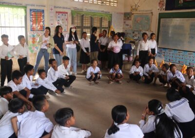 A group of students and teachers are gathered in a classroom, with some seated on the floor in a circle and others standing. The room, filled with educational posters and a whiteboard, buzzes with the spirit of the One Book One Love Campaign, reflecting an atmosphere of empowerment.
