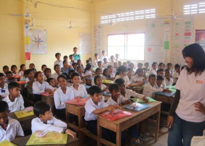 A teacher stands smiling at the front of a classroom full of students who are seated at their desks while some are standing. The classroom walls, adorned with various charts and posters, promote the One Book One Love Campaign that empowers families in Cambodia through reading initiatives.