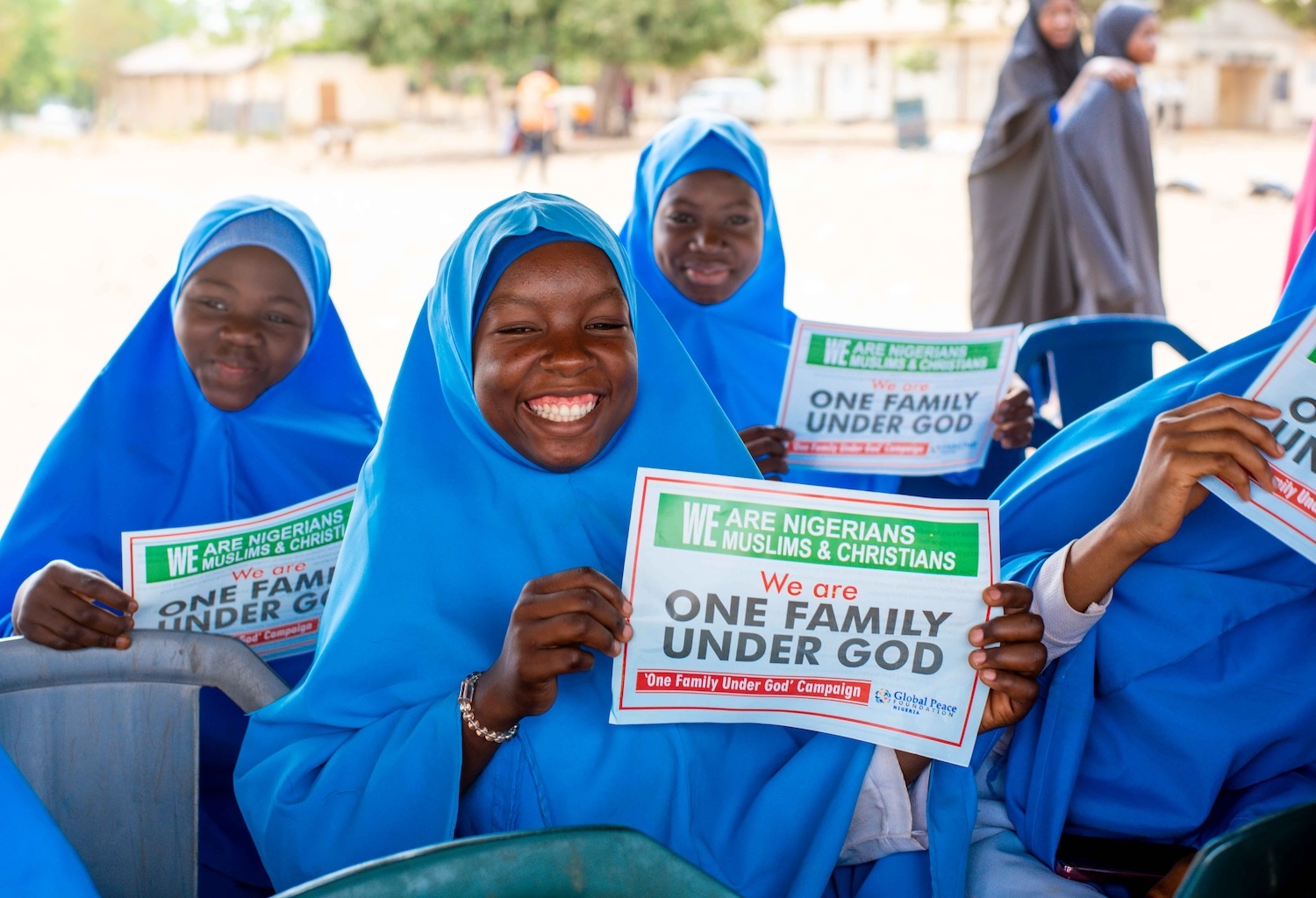 Group of smiling young students in blue hijabs holding signs promoting peacebuilding.