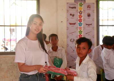 A woman in a white shirt hands a wrapped gift to a boy in a classroom setting with other children standing nearby. The scene, part of the One Book One Love Campaign, features educational posters on the wall and aims to empower families through literacy initiatives.