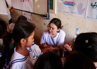 A woman in a white shirt sits and paints the hand of a child while other children watch attentively in a classroom setting. Posters with educational content adorn the walls, promoting the One Book One Love annual campaign.