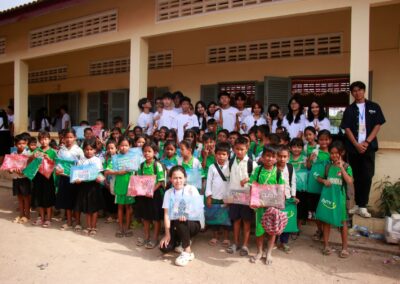 A large group of schoolchildren and adults pose for a photo in front of a school building, celebrating the One Book One Love Campaign. The children hold up colorful gift boxes, while some adults wear white shirts and some students wear green uniforms. Families in Cambodia join in the joyful occasion.