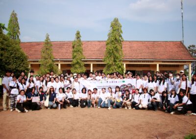 A large group of people, many wearing white shirts, pose outdoors in front of a building with trees. They hold a banner for the One Book One Love Campaign and appear to be participating in an annual gathering.