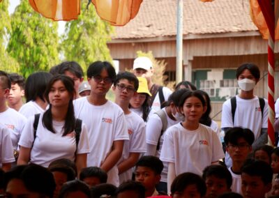 A group of students in white shirts, some wearing face masks, stand and sit under a shaded area with orange fabric above. They're outside near a building, participating in the One Book One Love Campaign that supports families in Cambodia.