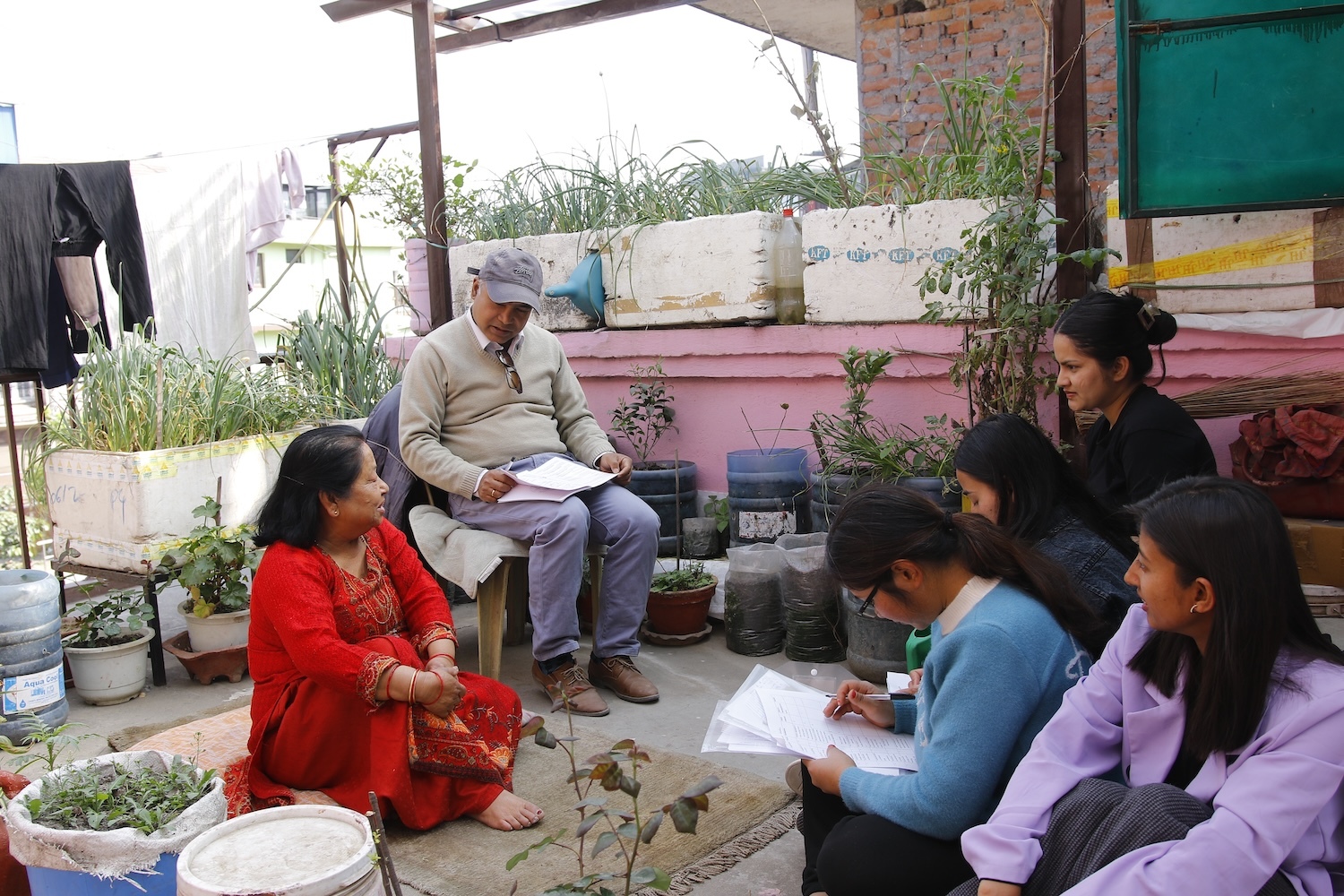 A group of people, including an elderly man and a woman and four younger women, sitting outdoors having a discussion with notebooks on their laps.