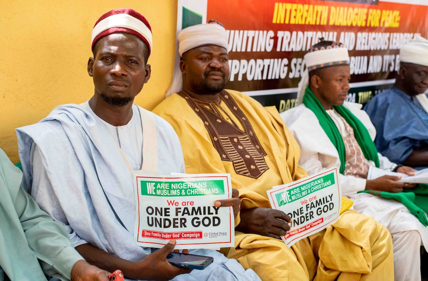 A group of men dressed in traditional attire hold signs reading "We are Nigerians, Muslims & Christians, We are one family under God" at an interfaith Community Dialogue event in Abuja aimed at promoting sustainable peace.