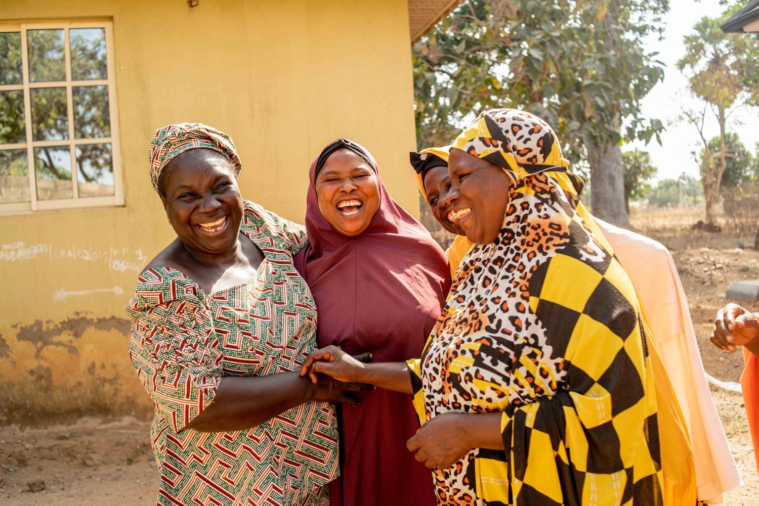 Four women, wearing colorful traditional attire, stand outside a building in Kagoro Chiefdom and laugh together while holding hands. Trees and a window are visible in the background, symbolizing the community resilience fostered through their interfaith dialogue.