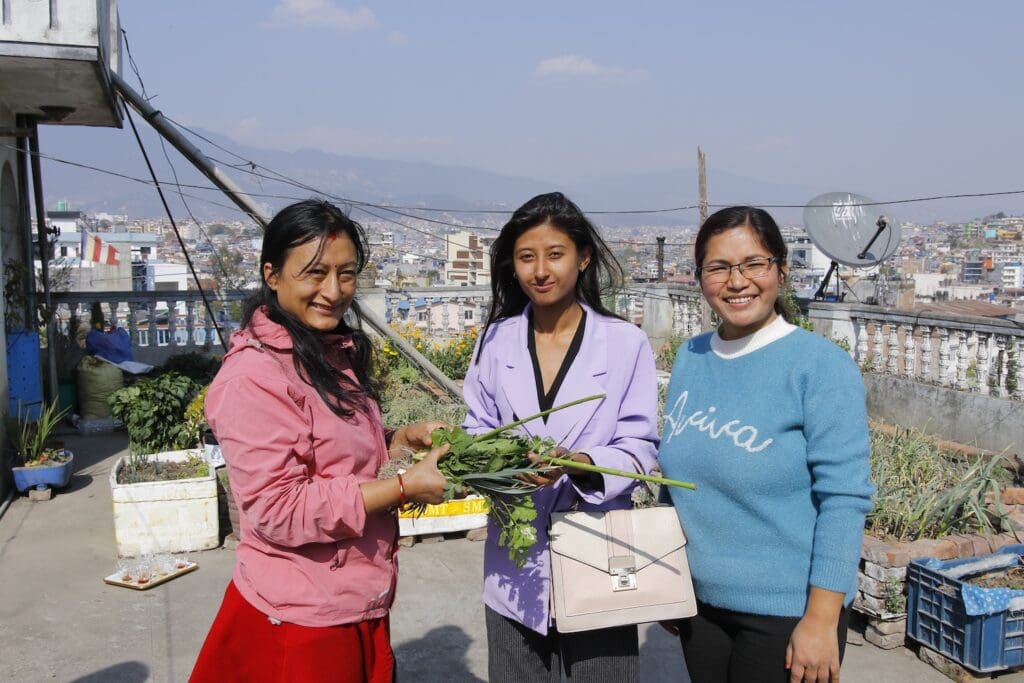 Three women holding plants on a rooftop garden as part of the Green Kathmandu Project, with the city skyline in the background.
