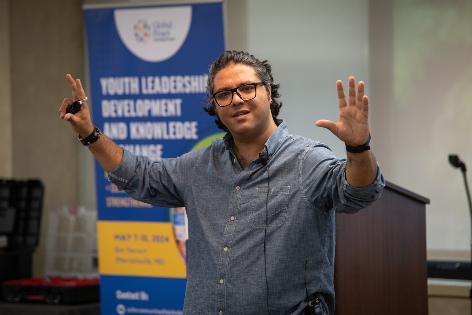 A person wearing glasses and a blue shirt is gesturing while speaking in front of a banner that reads "Youth Leadership Development and Knowledge Exchange," inspiring future youth leaders.