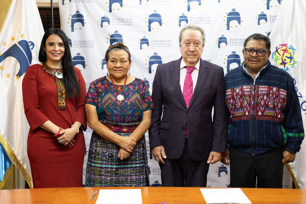 Four individuals are standing side by side, posing for a photo in front of a branded backdrop. Nobel Laureate Rigoberta Menchú and Former President Cerezo, both dressed in suits and glasses, stand alongside two women wearing traditional and modern attire, symbolizing peace in Central America.