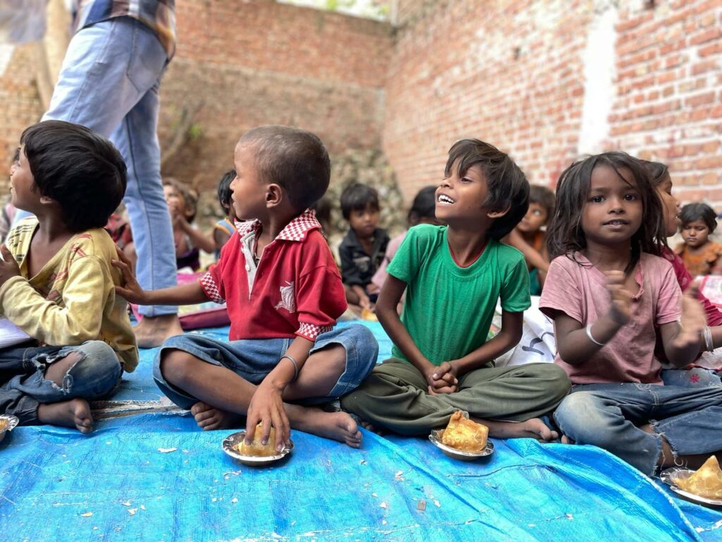 A group of children sitting on a blue sheet outdoors, eating food from small plates provided by volunteers. They are in an area surrounded by brick walls, part of a vibrant learning center in India.