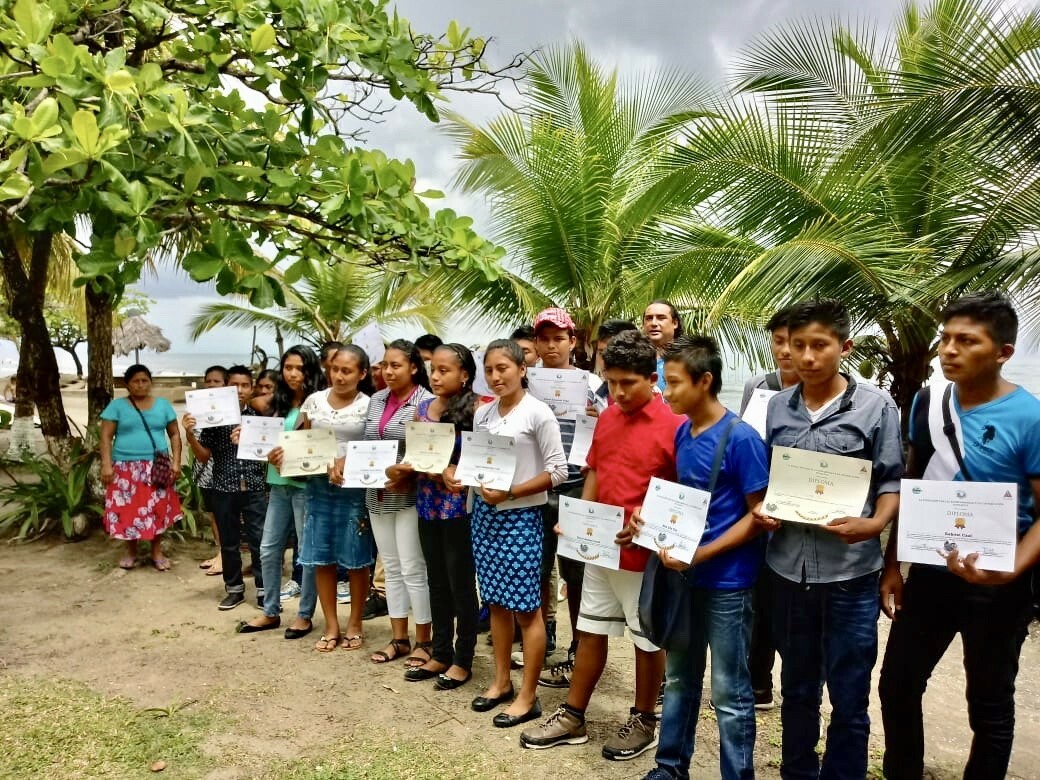 A group of young students, standing outdoors in Guatemala, proudly displaying their certificates with tropical trees in the background.