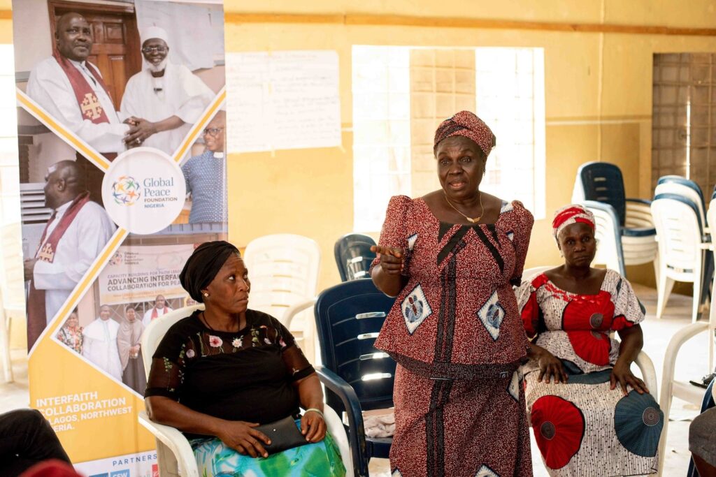 Three women are sitting and standing in a room; one woman is speaking. There is a banner behind them promoting interfaith dialogue and global peace. Several empty chairs are visible in the background, suggesting the event's focus on fostering community resilience within the Kagoro Chiefdom.