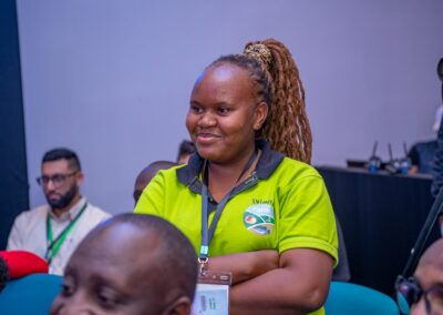 A person stands in a room, smiling and wearing a green polo shirt with a logo that reads "Iviani Farm." Other individuals are seated around them, discussing Environmental Track initiatives for GPLC Africa 2024. In the background, a person with a camera captures the moment.