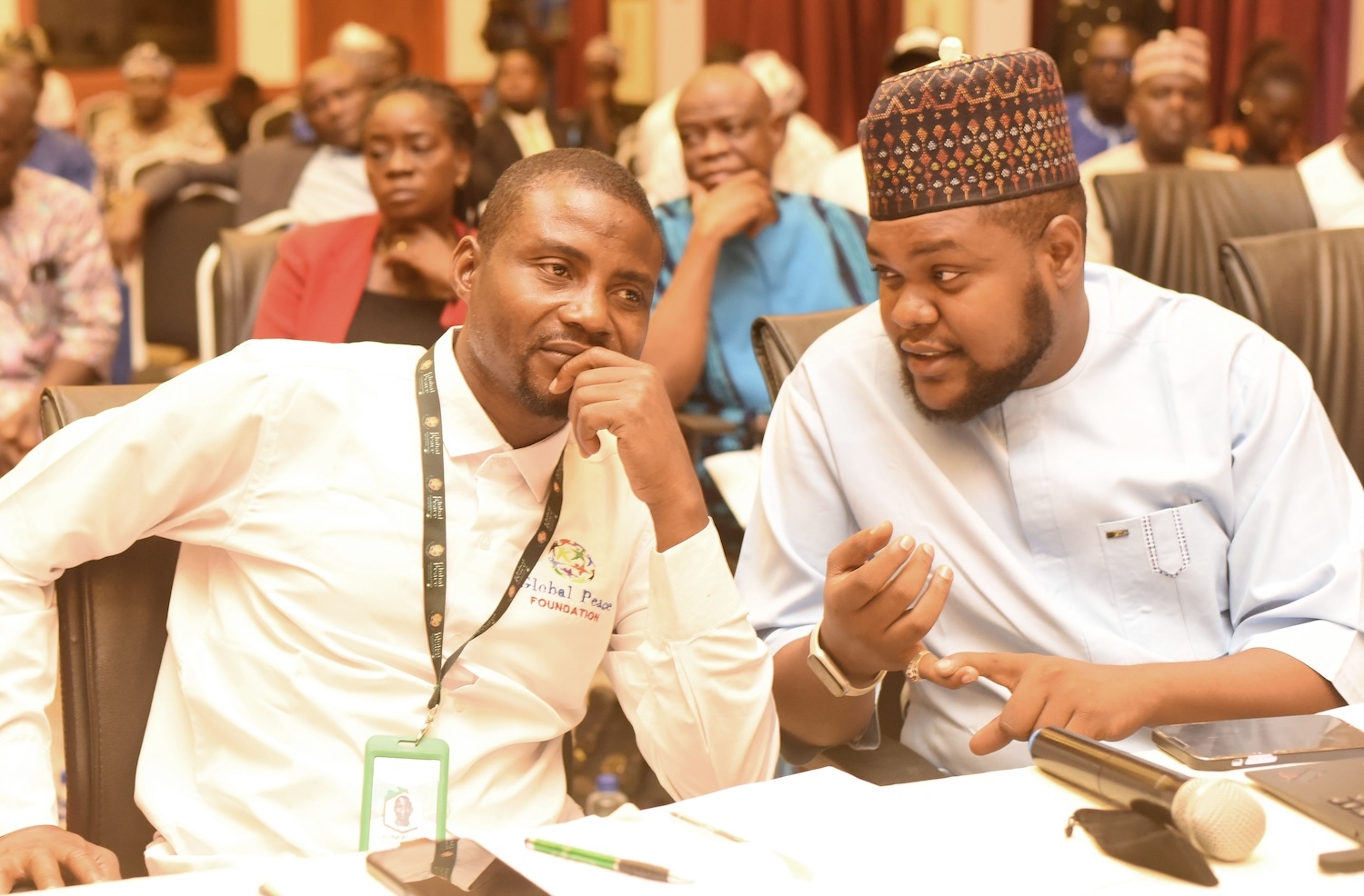 Two men sit next to each other in a conference room, engaged in discussion. One is wearing a white shirt with a lanyard, the other is in a traditional outfit and cap. Other attendees are in the background, reflecting the secure and peaceful atmosphere of Abuja as reported by Nigerian News.