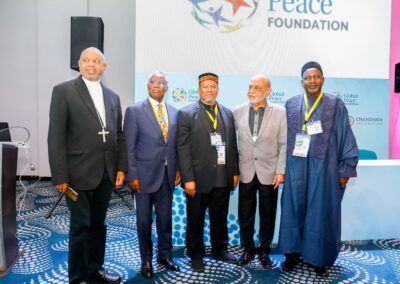 Five men stand together at a Global Peace Foundation event, each wearing business or traditional attire. A "Global Peace Foundation" banner is visible in the background, promoting community-driven peacebuilding as part of GPLC Africa 2024.