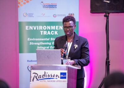 A person stands at a podium giving a presentation on environmental strategies at the Global Peace Leadership Conference. A banner in the background displays the event details and logos for GPLC Africa 2024's Environmental Track.