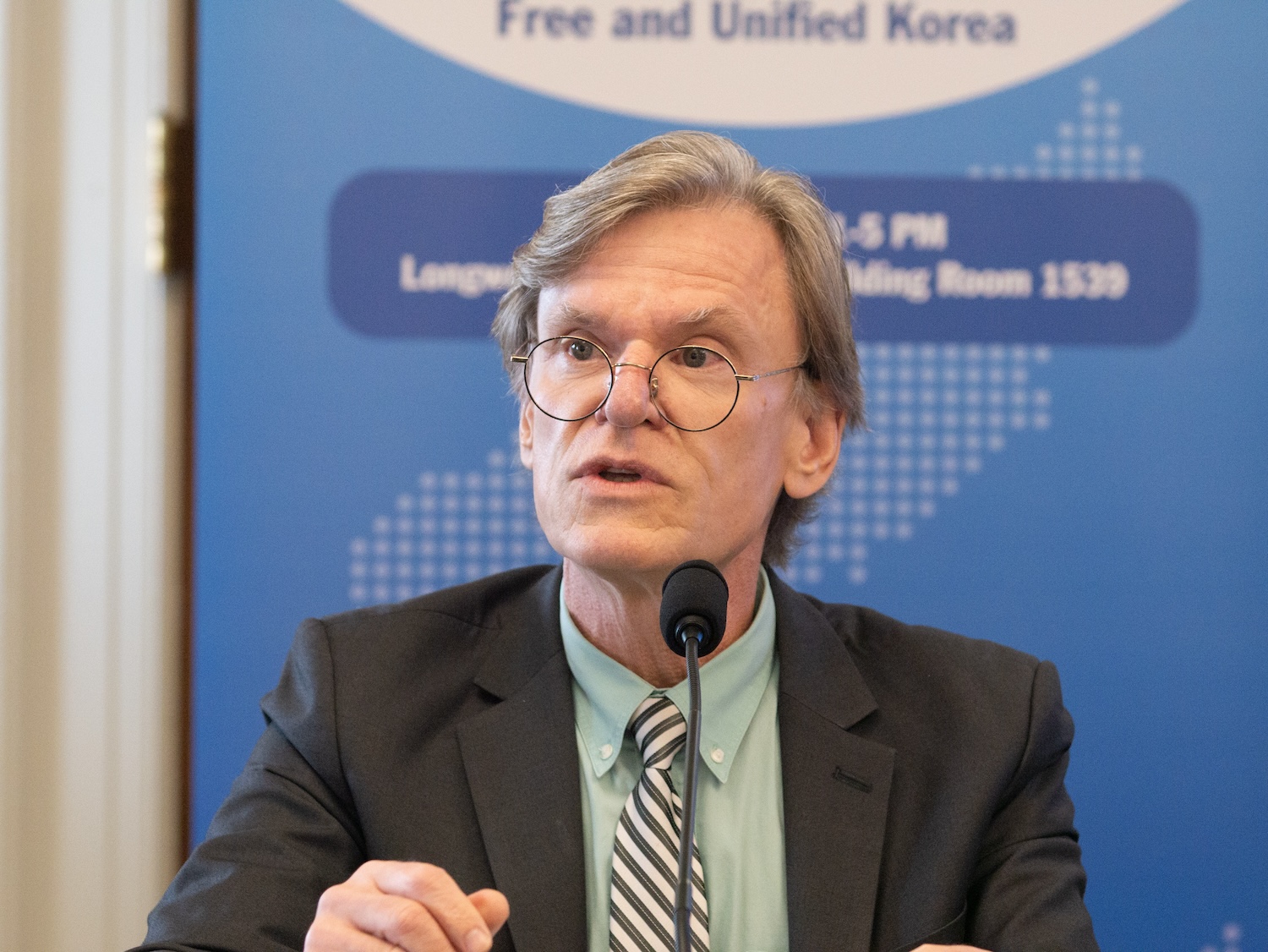 At the International Forum on One Korea, a man with glasses and gray hair speaks into a microphone. With a blue banner in the background, he stands confidently in his dark suit, green shirt, and striped tie advocating for U.S. support for a unified Korea.