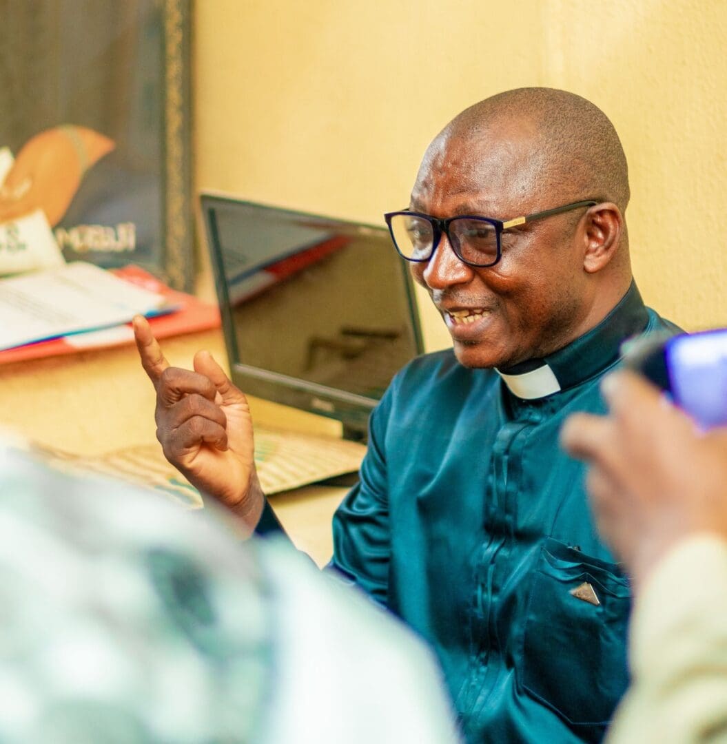 A man wearing glasses and clerical attire gestures while speaking, advocating for Social Cohesion, with a laptop and papers in the background. Another person is partially visible in the foreground.