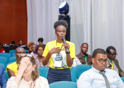 A woman in a yellow shirt stands at the microphone, addressing a packed auditorium filled with people seated on blue chairs during the GPLC Africa 2024 event, emphasizing the importance of community-driven peacebuilding.