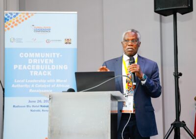 An individual stands at a podium speaking into a microphone. Behind them is a banner displaying information about the Global Peace Leadership Conference, held on June 26, 2023, in Nairobi, Kenya. Their address underscores the importance of community-driven peacebuilding ahead of GPLC Africa 2024.