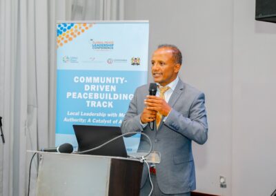 A man in a gray suit holding a microphone speaks at a podium with a "Community-Driven Peacebuilding Track" conference banner by GPLC Africa in the background.