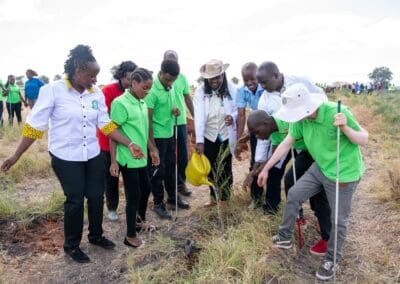 A group of people, some wearing green shirts and others in white or casual wear, gather around a planted sapling in an open field during the Kenya National Tree Growing Campaign organized by GPLC Africa.