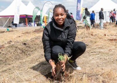 A person in a black jacket kneels on dry grass while planting a small tree sapling during the 2024 Kenya National Tree Growing Campaign. Tents and several people are visible in the background, highlighting the collaborative efforts spearheaded by GPLC Africa.