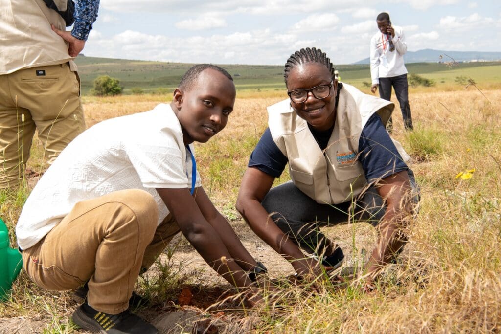 Two individuals are kneeling on the ground in a grassy field, planting a sapling as part of the Kenya National Tree Growing Campaign. Others are visible in the background, engaging in similar activities, contributing to GPLC Africa 2024's environmental goals.