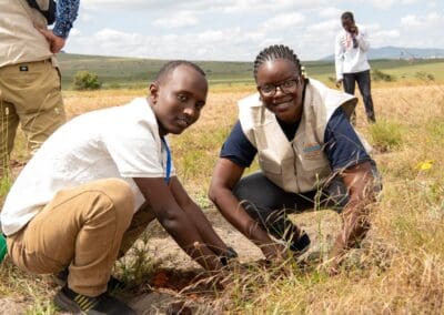 Two individuals are kneeling on the ground in a grassy field, planting a sapling as part of the Kenya National Tree Growing Campaign. Others are visible in the background, engaging in similar activities, contributing to GPLC Africa 2024's environmental goals.