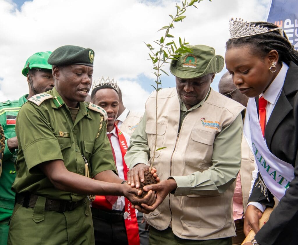 A group of people, including uniformed officials and individuals wearing sashes and crowns, collaborate in the Kenya National Tree Growing Campaign to plant a tree.