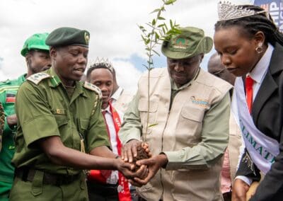 A group of people, including uniformed officials and individuals wearing sashes and crowns, collaborate in the Kenya National Tree Growing Campaign to plant a tree.