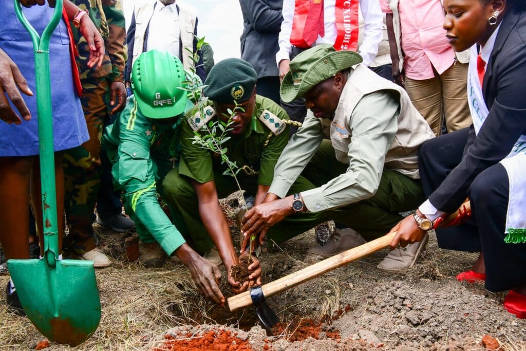 A group of people in uniforms and formal attire planting a small tree together, with shovels visible in the foreground and background, as part of the Kenya National Tree Growing Campaign linked to GPLC Africa 2024.
