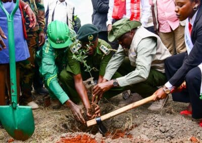 A group of people in uniforms and formal attire planting a small tree together, with shovels visible in the foreground and background, as part of the Kenya National Tree Growing Campaign linked to GPLC Africa 2024.