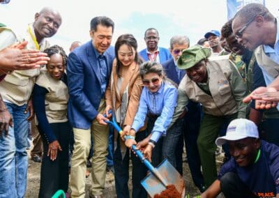 A group of people are gathered outdoors, participating in a tree-planting event hosted by GPLC Africa. Some individuals are holding a shovel with soil, while others look on and smile. This initiative is part of the Kenya National Tree Growing campaign for 2024.