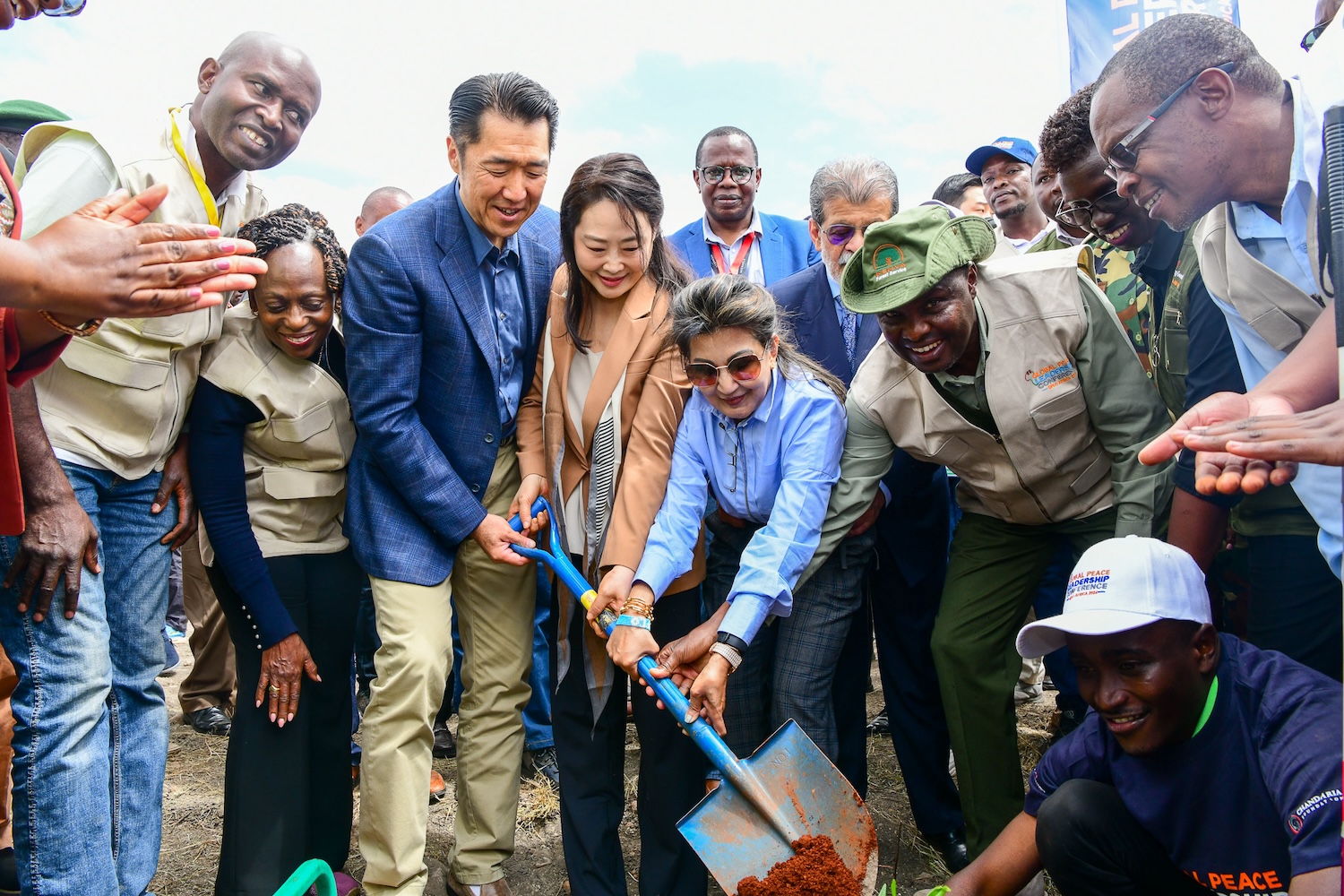 A group of people are gathered outdoors, participating in a tree-planting event hosted by GPLC Africa. Some individuals are holding a shovel with soil, while others look on and smile. This initiative is part of the Kenya National Tree Growing campaign for 2024.