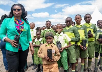 A woman stands next to a group of children in green-and-yellow school uniforms; they are outdoors and holding small potted plants, participating in the Kenya National Tree Growing campaign, with a cloudy sky in the background.