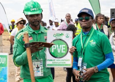 Two men stand in the foreground; one in a green uniform and helmet holding a shovel, the other in a green polo shirt holding a "Public Voluntary Education" sign. Behind them, a group of people is gathered for the Kenya National Tree Growing event hosted by GPLC Africa 2024.