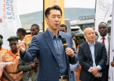A man in a blue blazer speaks into a microphone at an outdoor event, with a group of people, including some in scout uniforms, standing behind him, promoting the Kenya National Tree Growing Campaign.