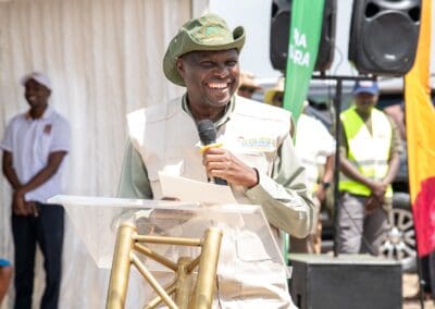 A man wearing a hat and vest speaks into a microphone at a podium during the GPLC Africa 2024 event. People in the background stand near a speaker and colorful banners promoting the Kenya National Tree Growing Campaign.