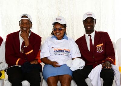Three people wearing uniforms and caps, with "Global Peace Leadership Conference" t-shirts, are seated and smiling while facing the camera. The backdrop is plain white. They are part of the 2024 Campaign for GPLC Africa focusing on Kenya National Tree Growing.
