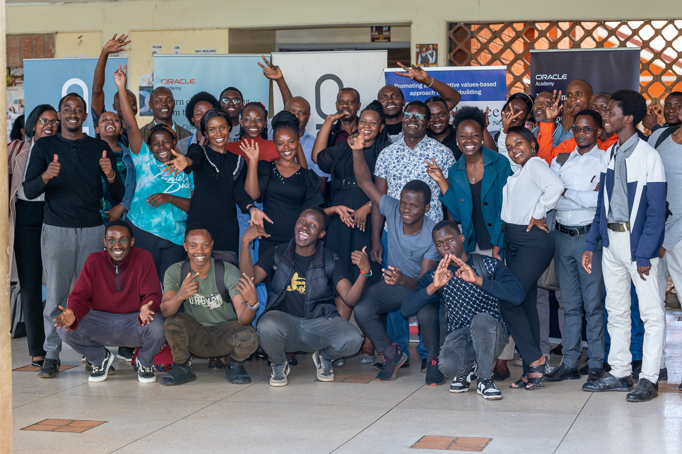 A group of people, some sitting and some standing, are posing for a group photo, many with smiles and cheerful gestures. They are indoors with banners from the Global Peace Foundation Kenya and Oracle Academy in the background, along with a tiled floor.