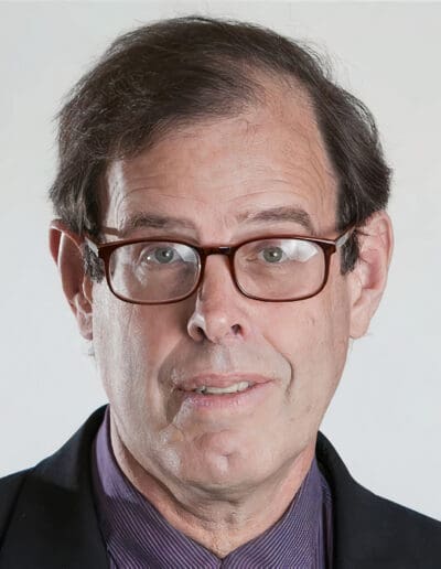 A man with glasses, short hair, and wearing a dark jacket over a purple shirt, looks directly at the camera against a plain background.
